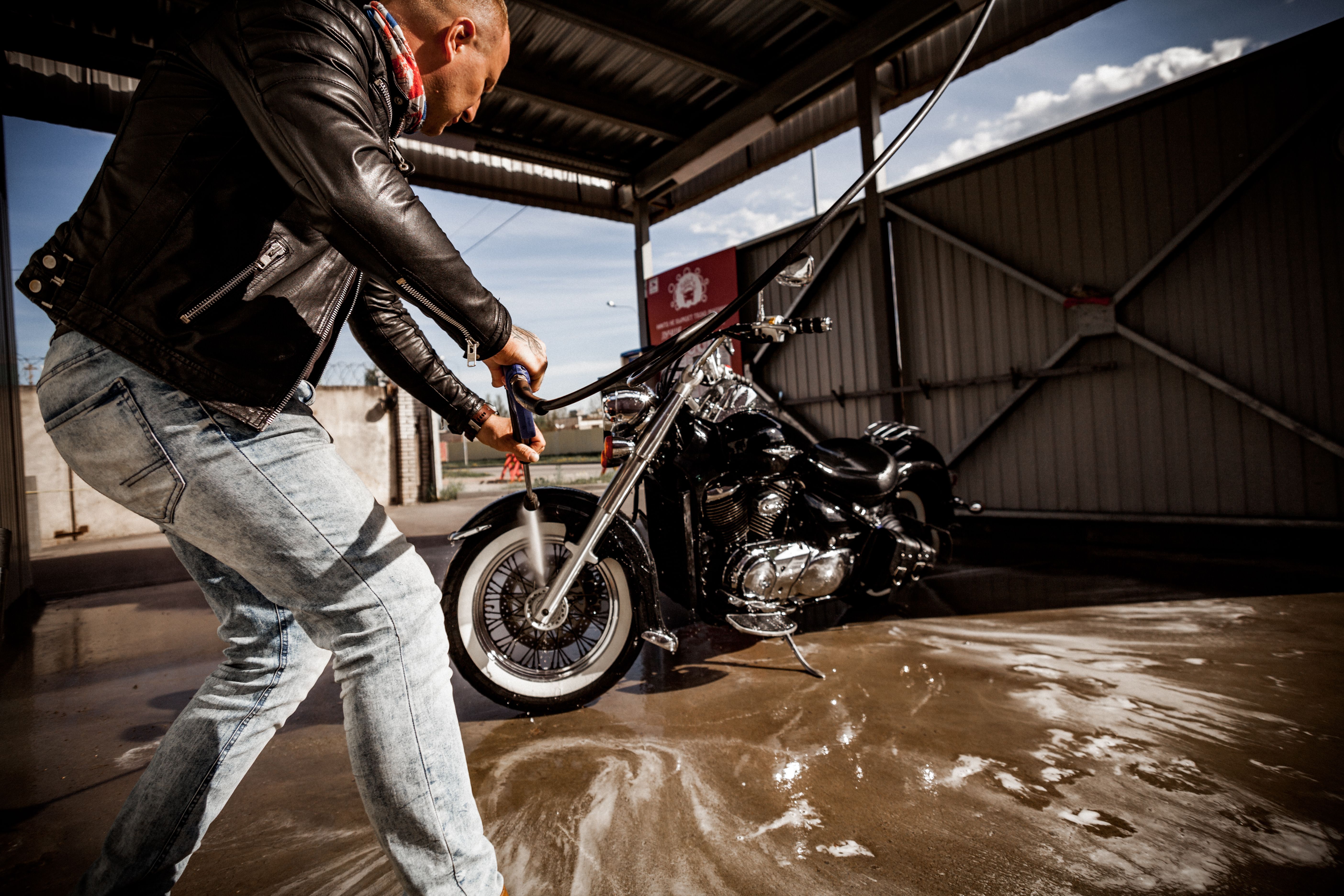 Man washing a motorcycle with a hose at an outdoor wash station on a sunny day